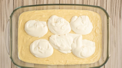 A glass baking dish with a layer of yellow Lemon Coffee Cake batter, topped with six dollops of white creamy mixture, sits on a light wooden surface.
