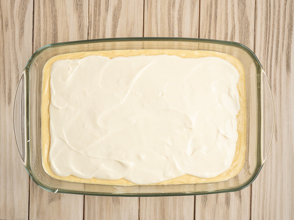 A glass baking dish filled with an unbaked Lemon Coffee Cake, featuring a smooth, creamy white layer spread evenly over a yellow dough base, set on a light wooden surface.