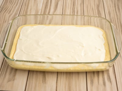 A glass baking dish contains an unbaked layered Lemon Coffee Cake dessert with a smooth white creamy layer on top, resting on a light wooden surface.
