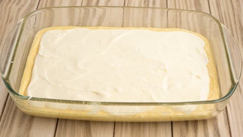 A glass baking dish contains an unbaked layered Lemon Coffee Cake dessert with a smooth white creamy layer on top, resting on a light wooden surface.