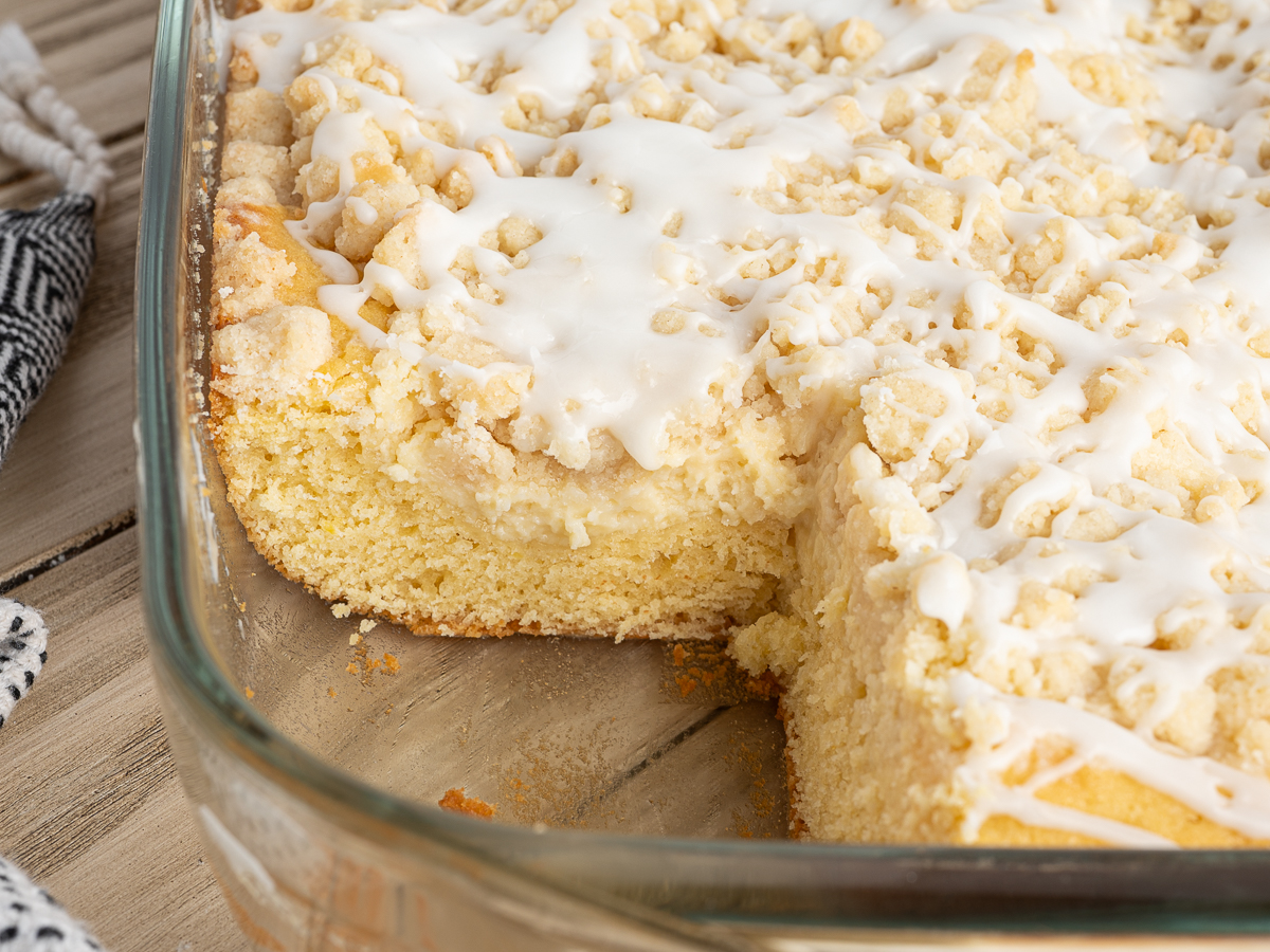 A glass baking dish filled with crumbly Lemon Coffee Cake topped with white icing. One piece has been cut and removed, revealing the soft, fluffy texture inside.