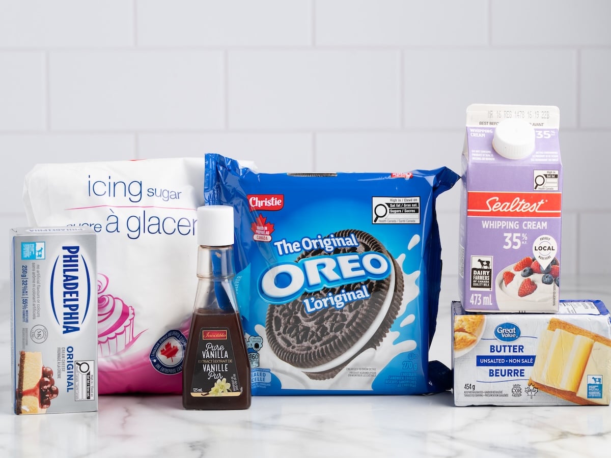 Ingredients for Oreo Frosting: a bag of icing sugar, a package of original Oreo cookies, Philadelphia cream cheese, vanilla extract, whipping cream, and unsalted butter on a marble countertop.