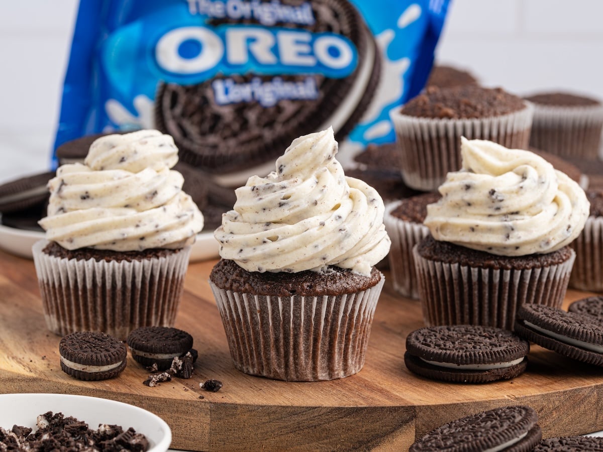 Three chocolate cupcakes with Oreo Frosting are displayed on a wooden board, surrounded by whole and crumbled Oreo cookies, with a blue Oreo package in the background.