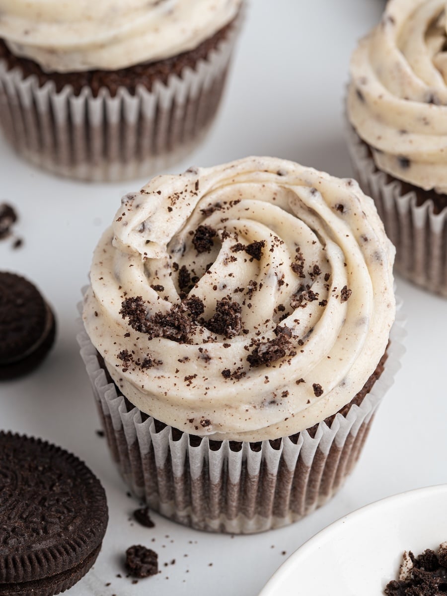 Chocolate cupcake topped with rich Oreo frosting and sprinkled with cookie crumbs, surrounded by sandwich cookies on a white surface.