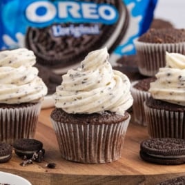 Chocolate cupcakes topped with swirled Oreo Frosting sit on a wooden board, surrounded by Oreo cookies and a blue Oreo package in the background.