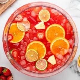 Large glass drink dispenser filled with bright red party punch, orange and lime slices, strawberries, and pineapple chunks surrounded by tropical fruit.