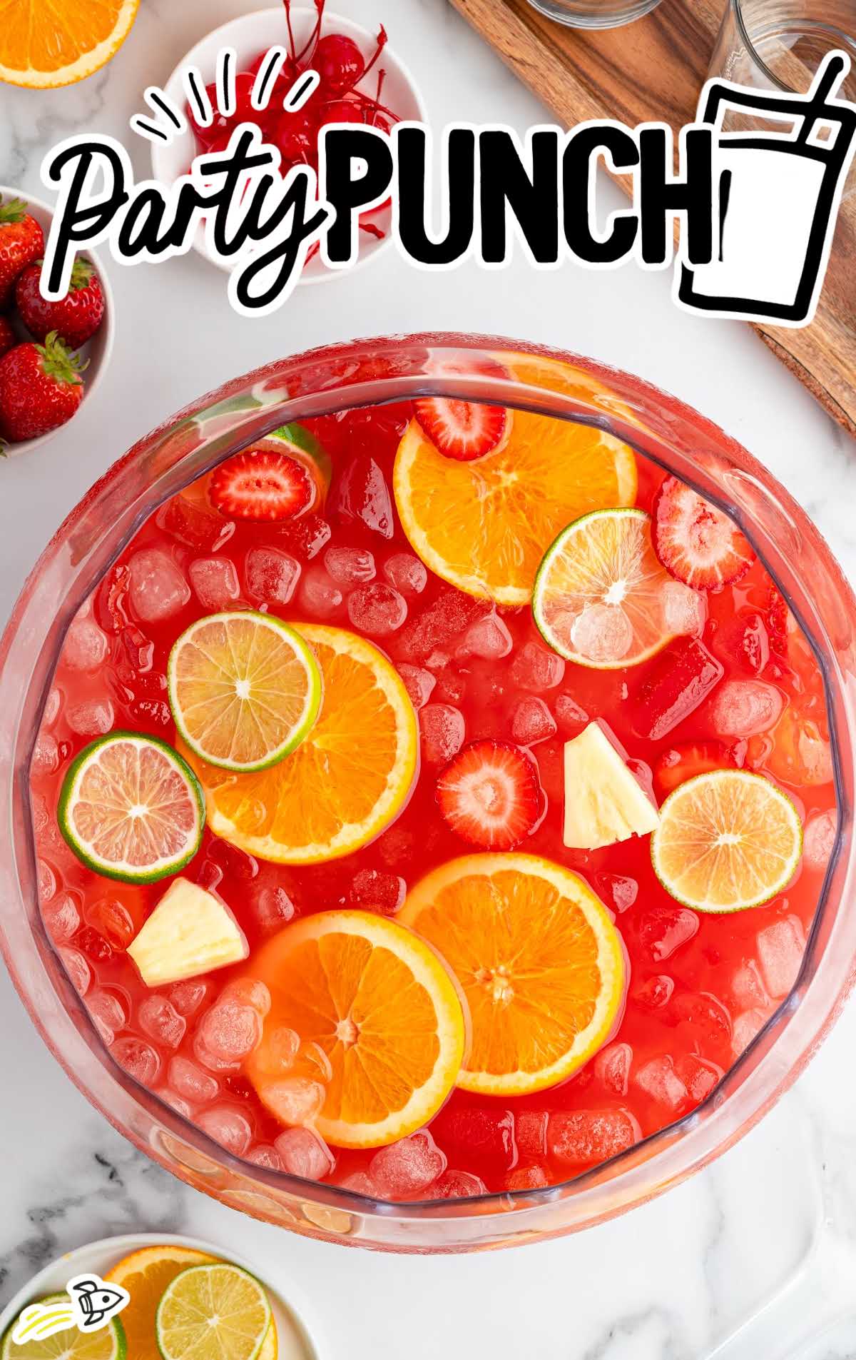 Overhead view of a punch bowl filled with ice, orange slices, lime slices, strawberries, and pineapple pieces for a refreshing tropical party punch.