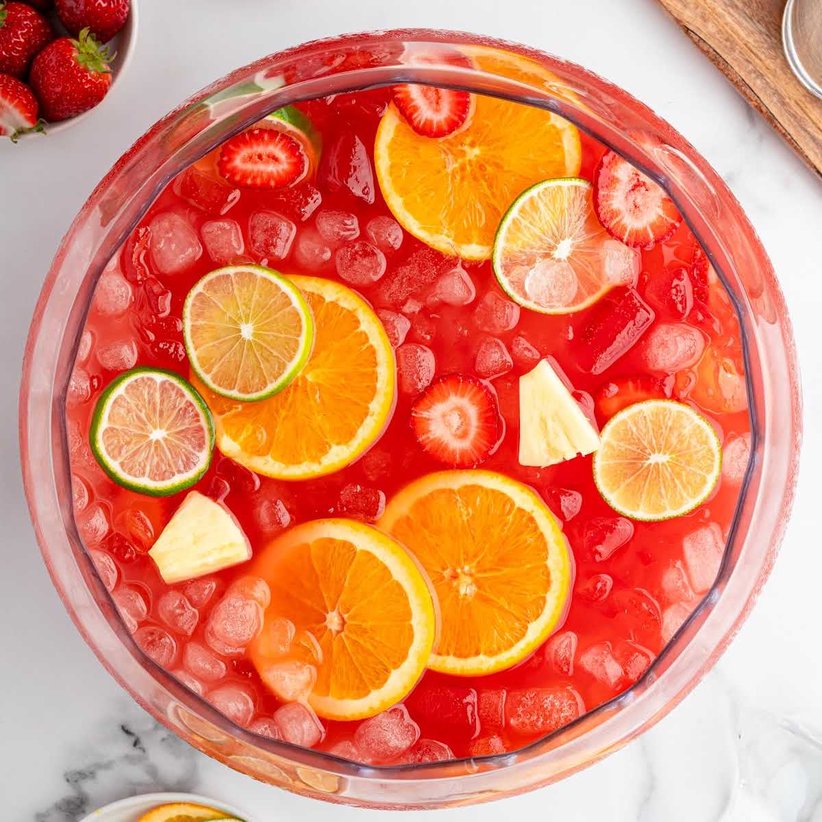Overhead view of red party punch in a clear bowl with citrus and strawberry garnish, styled for a recipe card layout.