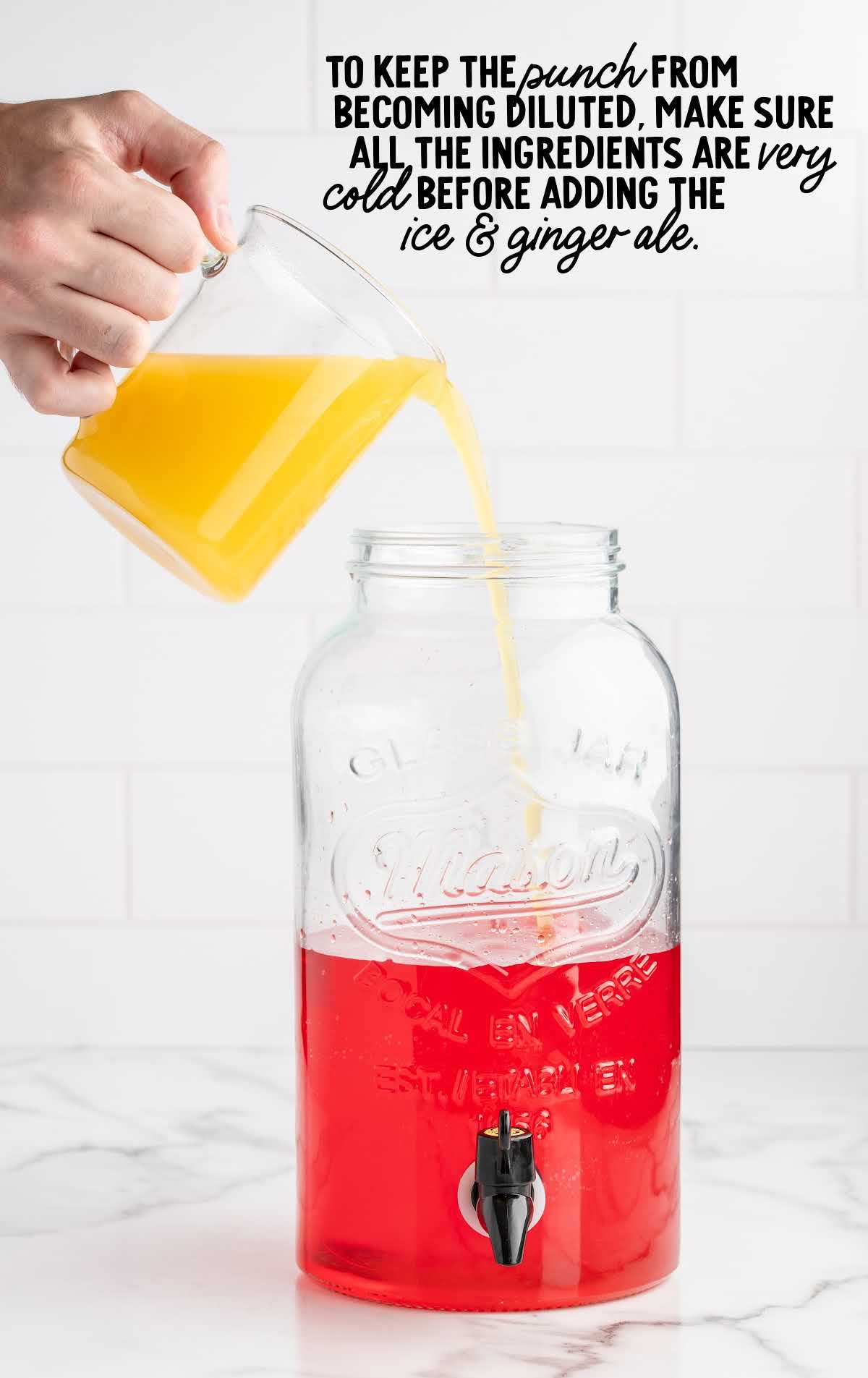 Step image showing chilled orange juice being poured into red Hawaiian Punch base in a mason jar dispenser for tropical party punch.