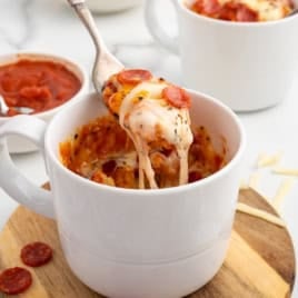 A fork lifts a cheesy, pepperoni-topped bite from a white mug filled with Pizza in a Mug, a pizza-like dish. Another mug and a bowl of sliced pepperoni are in the background on a wooden board.