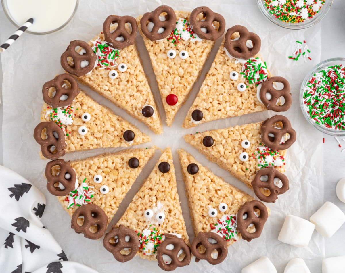 Overhead view of a full circle of reindeer-shaped Rice Krispie treat wedges decorated with chocolate-covered pretzel antlers, candy eyes, M&M noses, and Christmas sprinkles, arranged on parchment with bowls of sprinkles and glasses of milk.