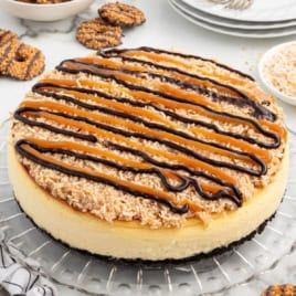 A Samoa Cheesecake with a chocolate cookie crust, topped with toasted coconut, caramel, and chocolate drizzle. Plates and forks are in the background, along with cookies and extra coconut.