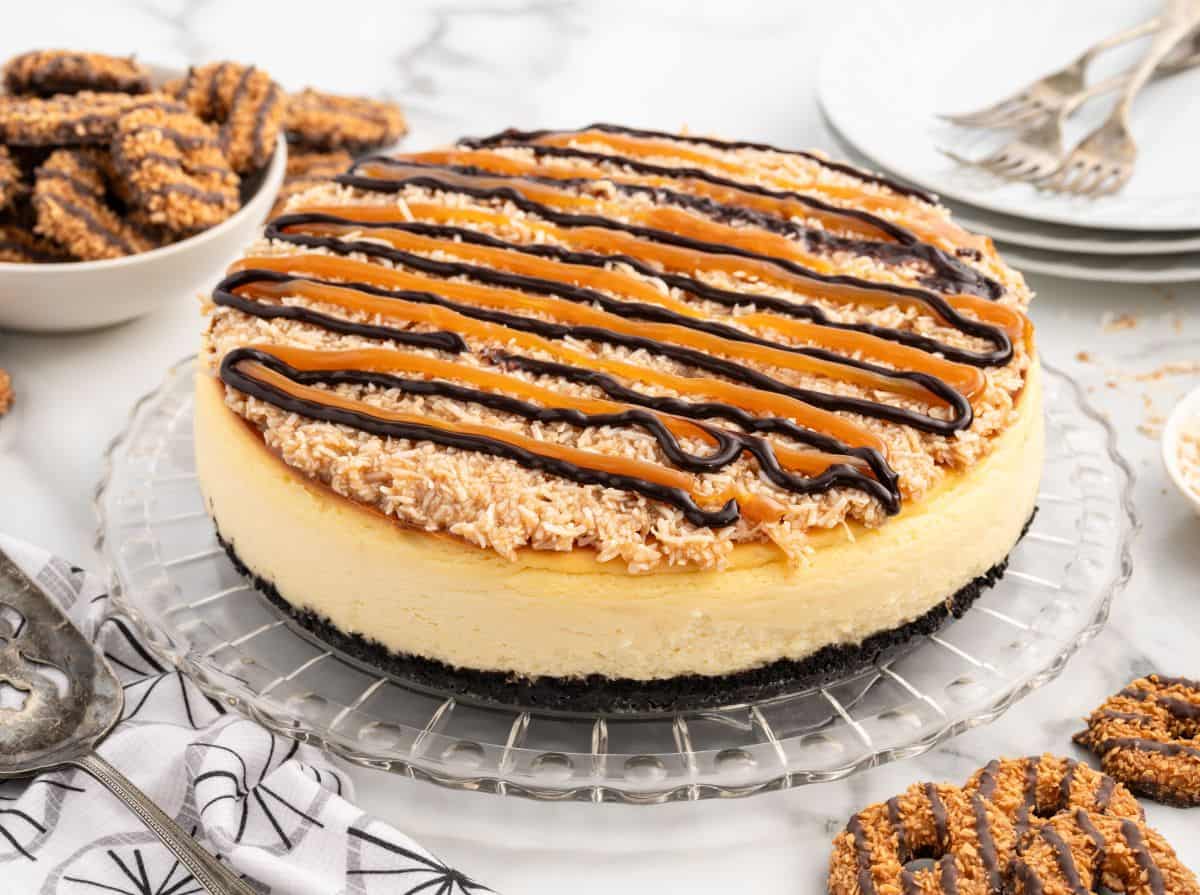 A Samoa Cheesecake with a chocolate cookie crust, topped with toasted coconut, caramel, and chocolate drizzle, sits on a glass platter. Plates, forks, and Samoas cookies are nearby on a marble surface.