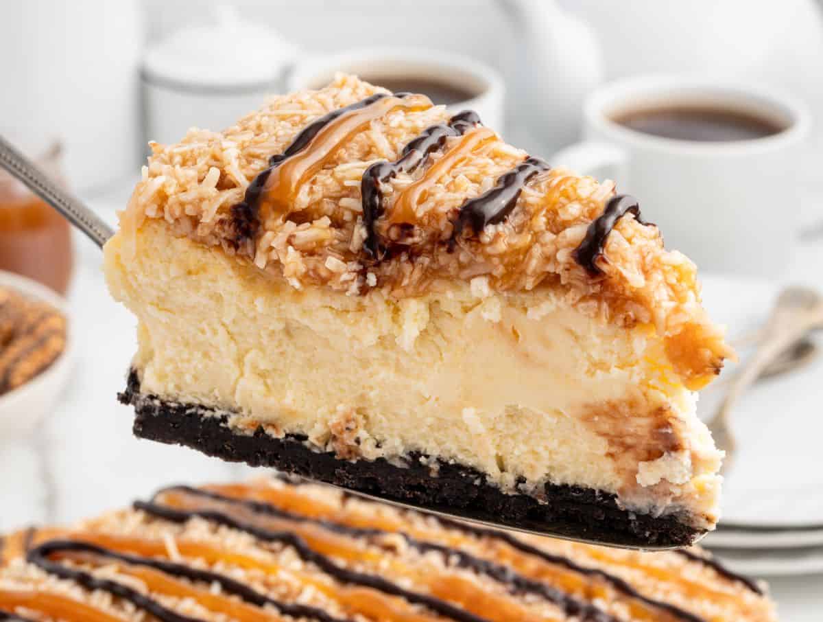 A close-up of a slice of Samoa Cheesecake with a chocolate cookie crust, topped with toasted coconut, caramel, and chocolate drizzle. Coffee cups and a cheesecake are blurred in the background.