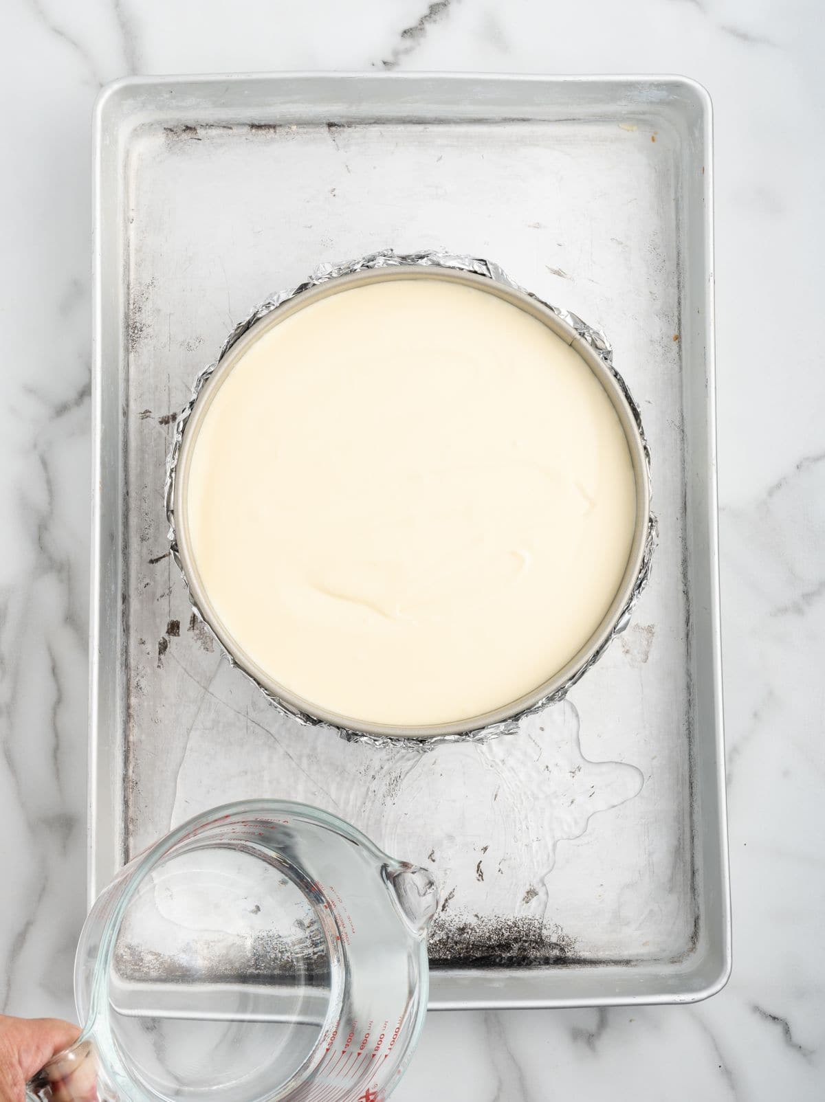 A hand pours water from a glass measuring cup into a baking sheet holding a Samoa Cheesecake in a round pan, preparing a water bath on a marble surface.