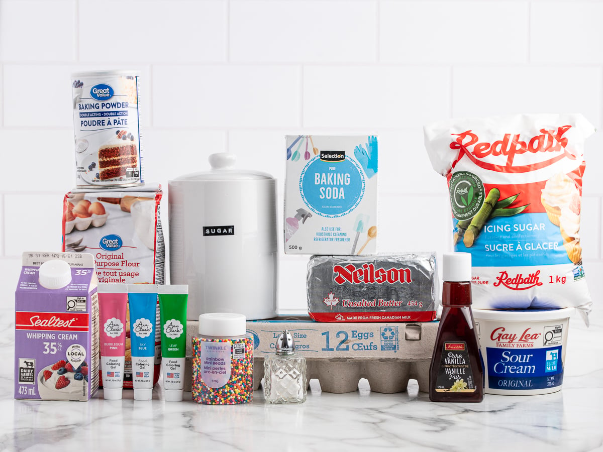 A variety of baking ingredients on a counter, perfect for making Sour Cream Cookies, including flour, sugar, baking powder, baking soda, icing sugar, eggs, butter, whipping cream, sour cream, vanilla, gel food colors, sprinkles, and a salt shaker.