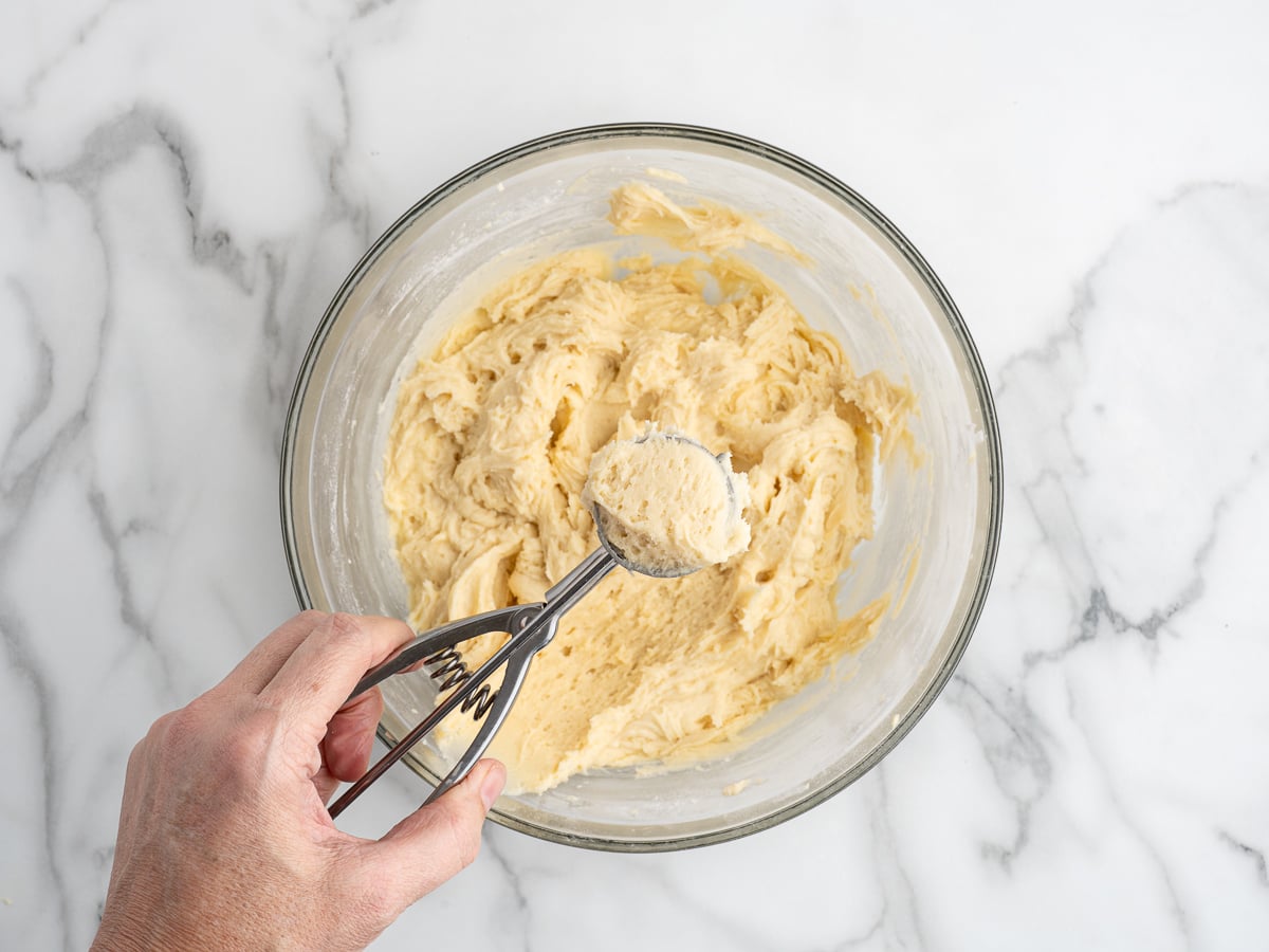 A hand holds a cookie scoop with raw dough above a glass bowl filled with creamy Sour Cream Cookies dough, set on a white marble surface.