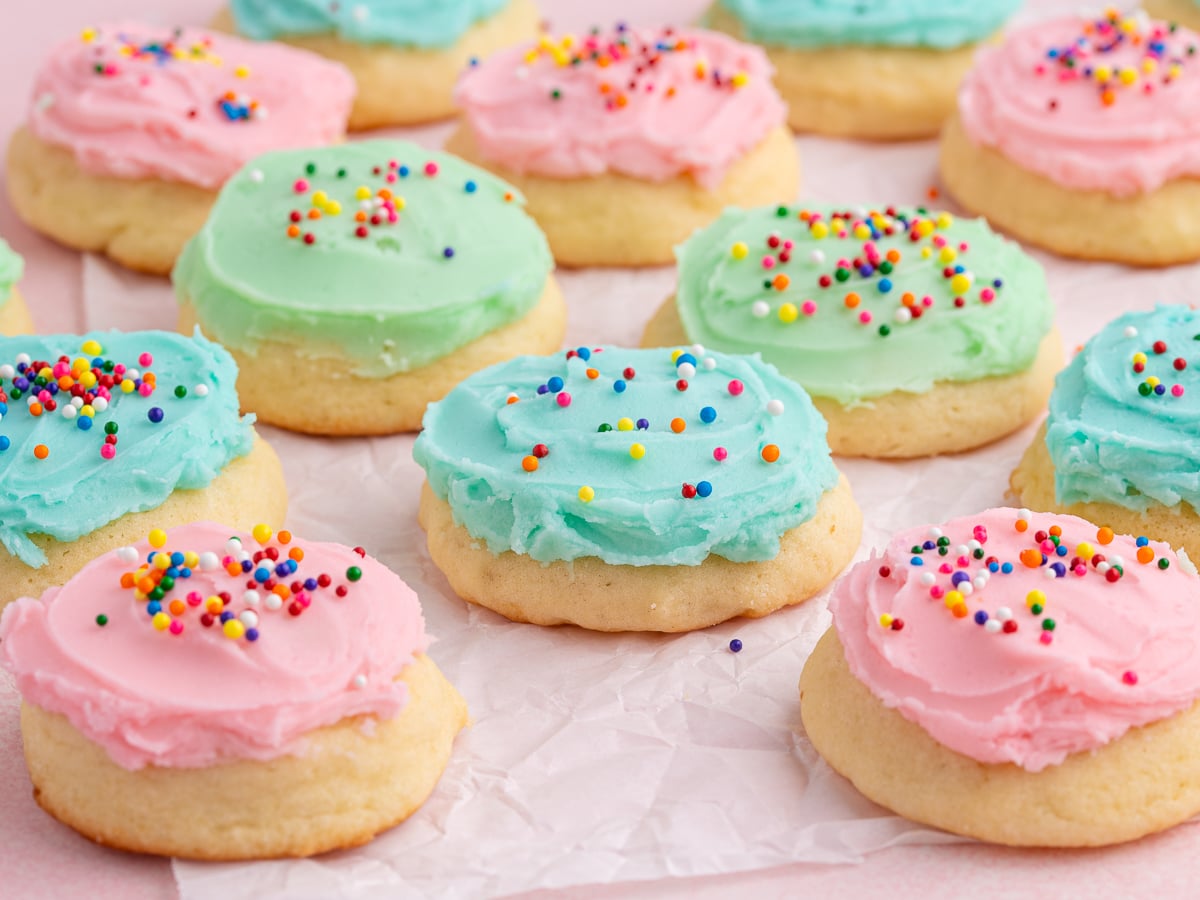 Sour Cream Cookies with pastel pink, blue, and green icing are arranged on crumpled parchment paper. Some frosted cookies are topped with colorful sprinkles.