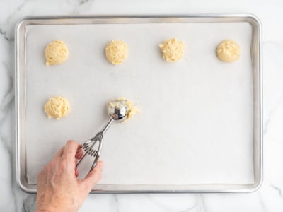 A hand uses a metal scoop to place balls of sour cream cookie dough on a parchment-lined baking sheet, with five dough balls already spaced out across the tray.