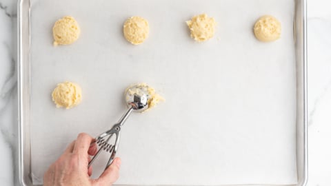 A hand uses a metal scoop to place balls of sour cream cookie dough on a parchment-lined baking sheet, with five dough balls already spaced out across the tray.