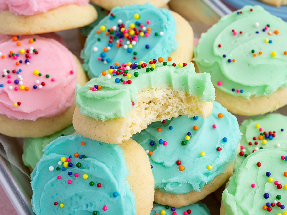 A tray of frosted Sour Cream Cookies with colorful icing in pink, blue, and green, topped with rainbow sprinkles. One green-iced cookie has a bite taken out of it, showing the soft, fluffy texture inside.