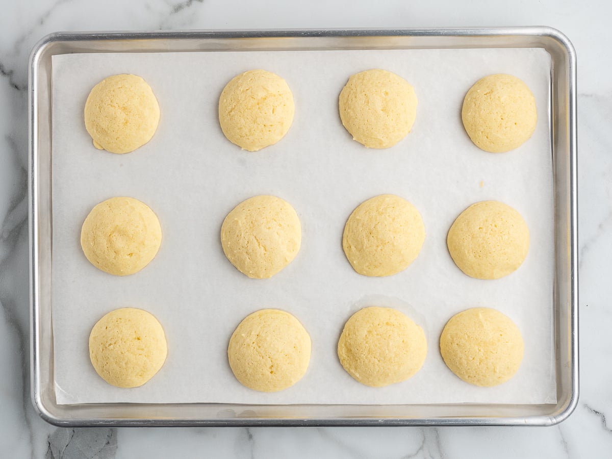 Twelve round, golden Sour Cream Cookies are evenly spaced on a parchment-lined baking sheet, resting on a marble surface.