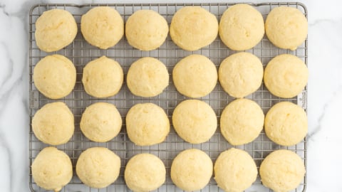 Twenty-four round, golden Sour Cream Cookies are arranged in a grid pattern on a cooling rack, set on a white marble surface.