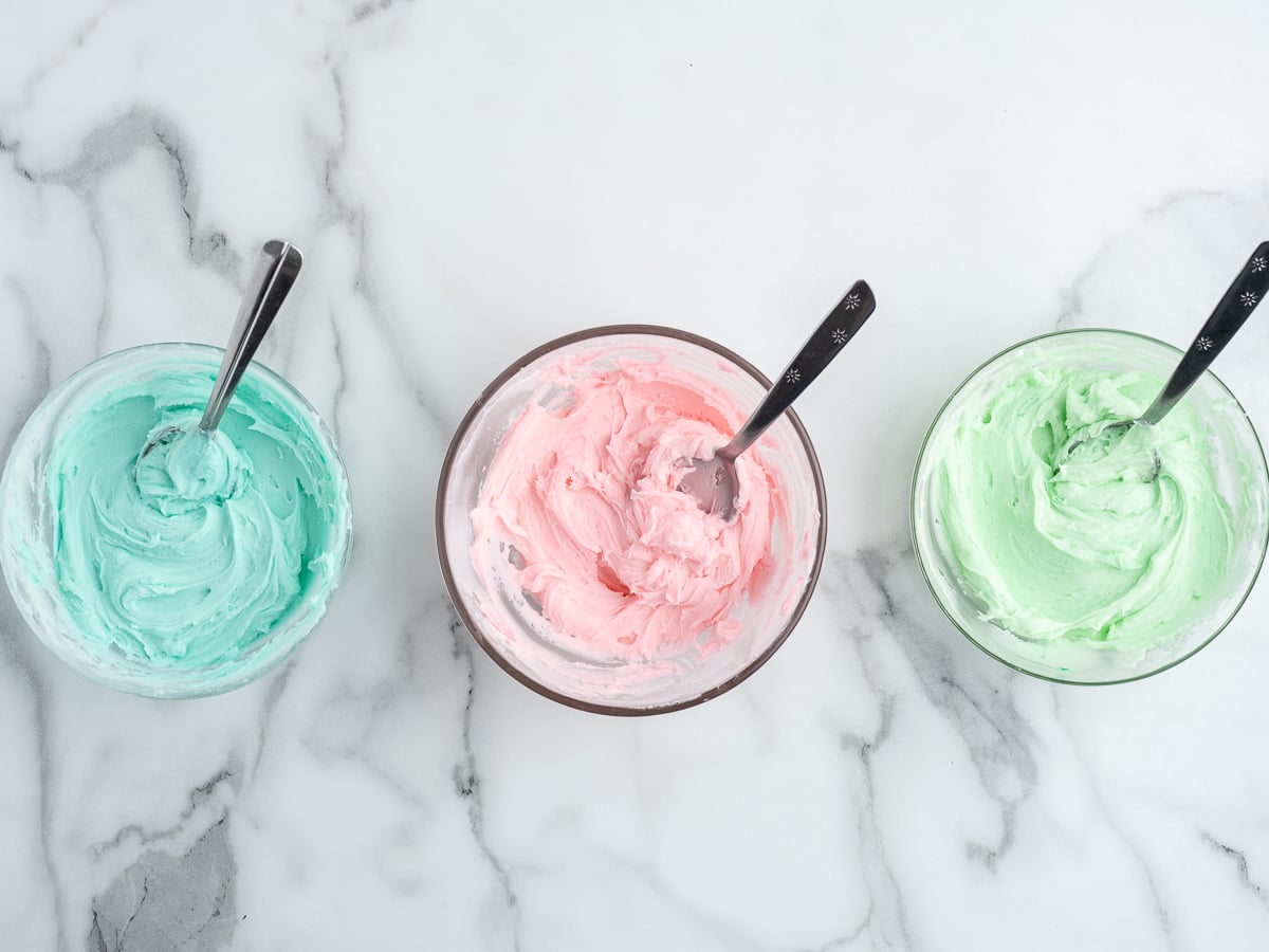 Three glass bowls with metal spoons, each filled with pastel-colored frosting—blue, pink, and green—are arranged on a white marble surface, ready to decorate freshly baked Sour Cream Cookies.