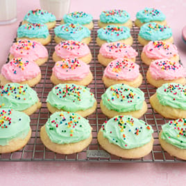 Rows of sour cream cookies with pink and green frosting and colorful sprinkles cool on a wire rack. Glasses of milk and plates sit in the background atop a cheerful pink surface.