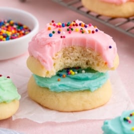 Two soft Sour Cream Cookies are stacked, one with blue frosting and the top one with pink frosting and rainbow sprinkles. The top cookie has a bite taken out. A bowl of sprinkles and more cookies are in the background.