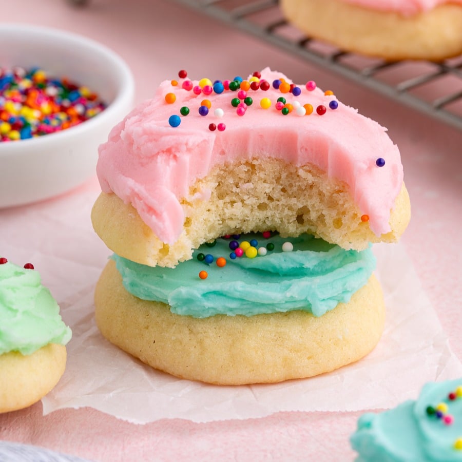 Two soft Sour Cream Cookies are stacked, one with blue frosting and the top one with pink frosting and rainbow sprinkles. The top cookie has a bite taken out. A bowl of sprinkles and more cookies are in the background.