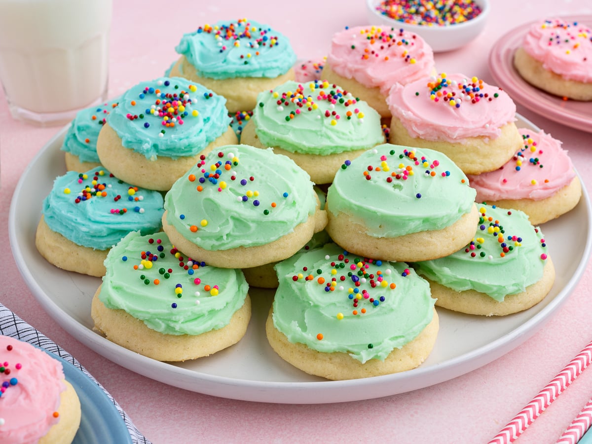 A plate of soft sour cream cookies topped with pink, blue, and green frosting, each decorated with colorful round sprinkles. Bowls of extra sprinkles and a glass of milk are in the background.