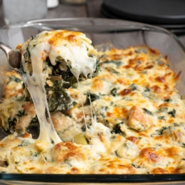 A spoon lifts a cheesy, baked Spinach and Artichoke Chicken Casserole from a glass dish, with melted cheese stretching from the serving. The casserole is golden brown on top and appears creamy and hearty.