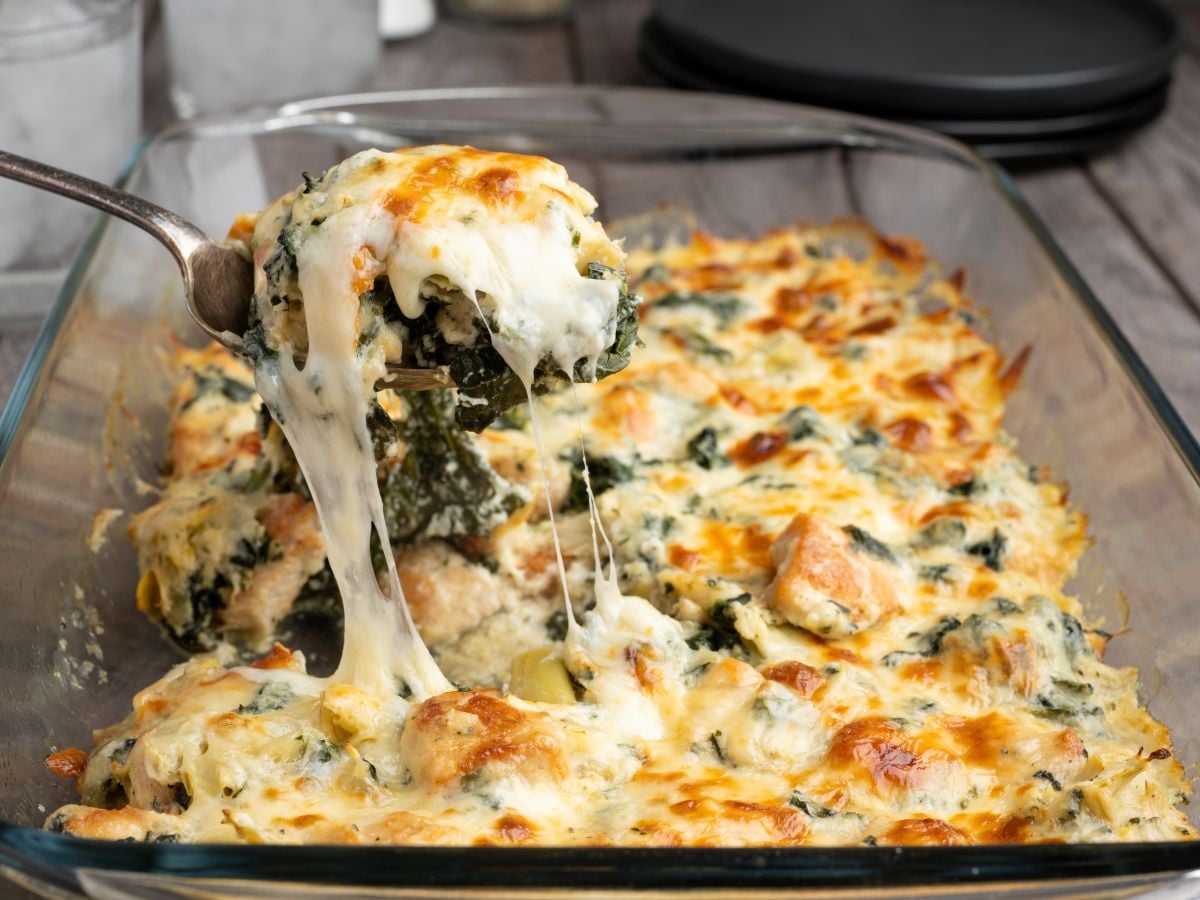 A spoon lifts a cheesy, baked Spinach and Artichoke Chicken Casserole from a glass dish, with melted cheese stretching from the serving. The casserole is golden brown on top and appears creamy and hearty.