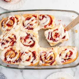 A glass baking dish with iced raspberry sweet rolls; one roll is being lifted out with a spatula. Reminiscent of Strawberry Cinnamon Rolls, the rolls are swirled with vibrant raspberry filling and topped with a generous layer of white icing.