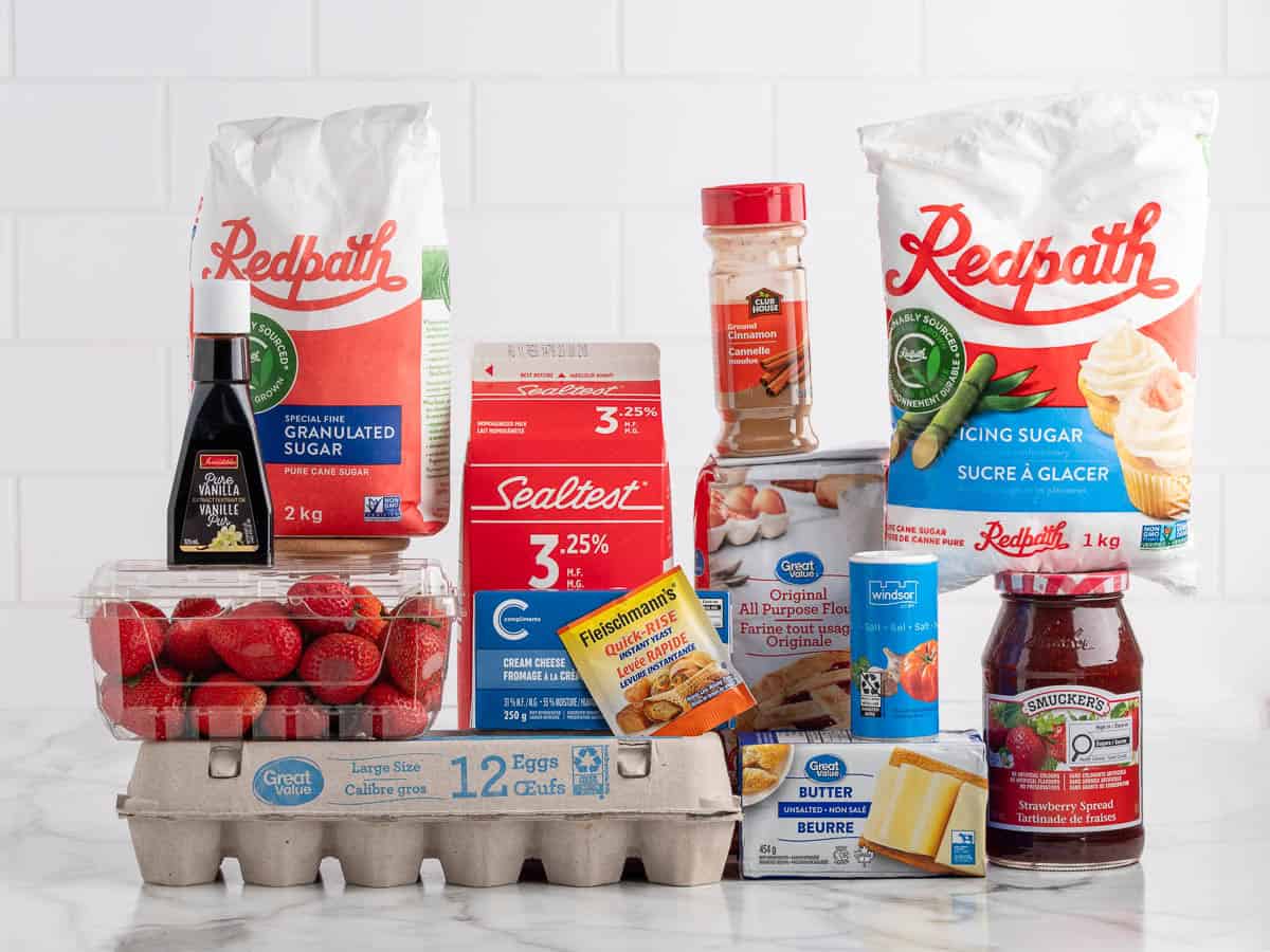 Groceries on a counter, including eggs, strawberries, butter, milk, sugar, icing sugar, vanilla, cinnamon, baking powder, cream cheese, and strawberry jam—everything you need to create delicious Strawberry Cinnamon Rolls—arranged in front of a white tiled wall.