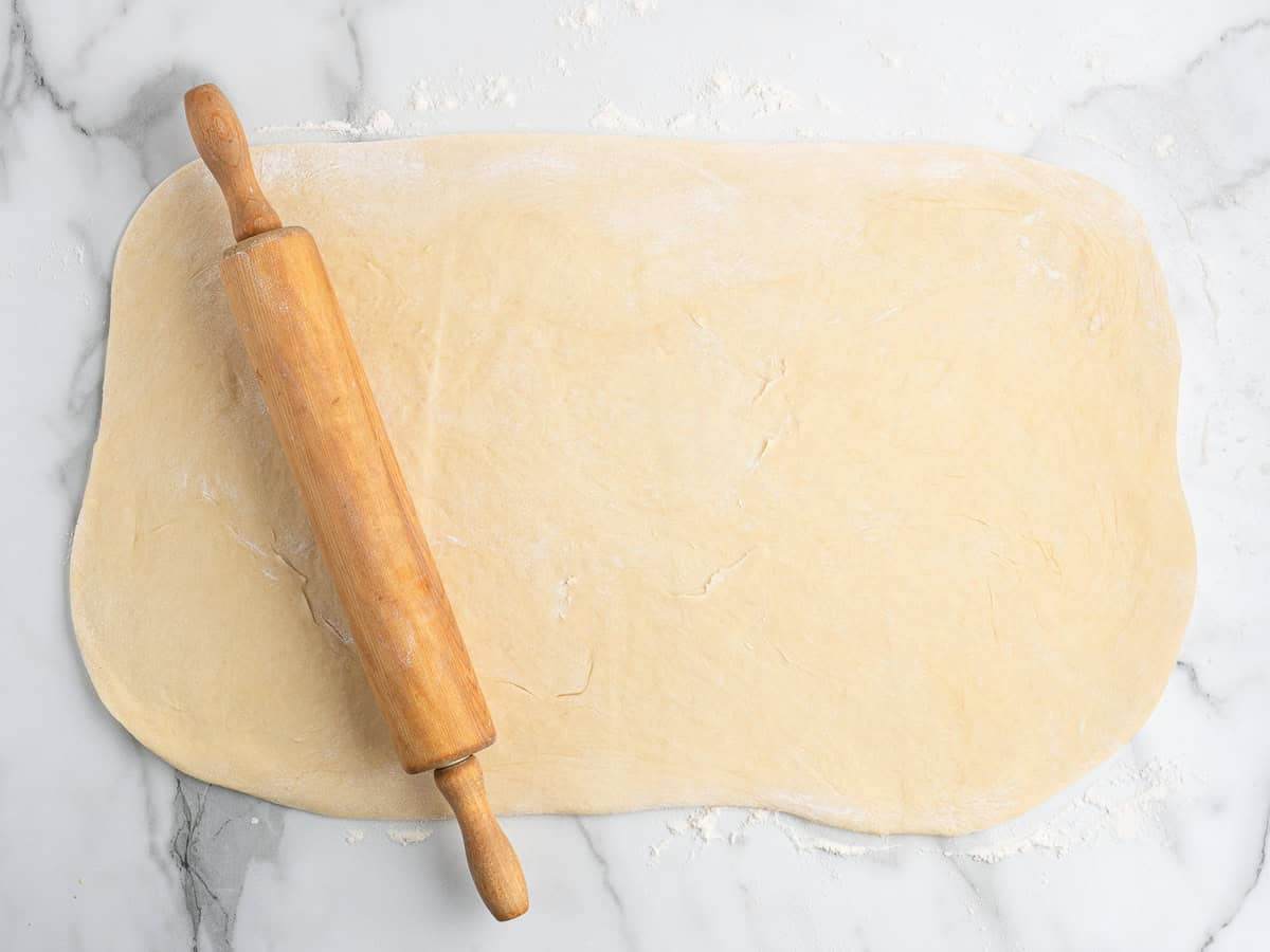 A wooden rolling pin rests on a sheet of rolled-out dough for Strawberry Cinnamon Rolls on a floured marble surface, ready for baking preparations.
