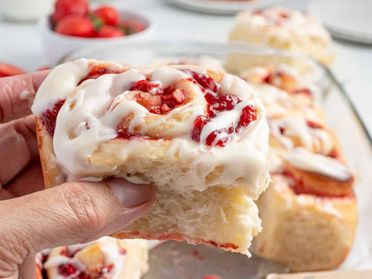 A hand holds a thick, fluffy strawberry cinnamon roll topped with white icing and strawberry filling. More strawberry cinnamon rolls and a bowl of strawberries are visible in the background.