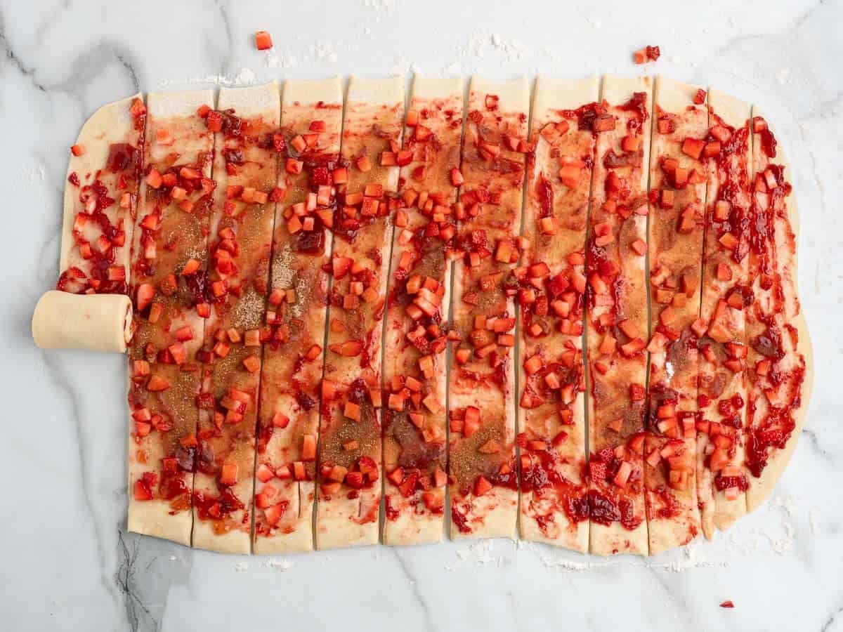 Rectangular dough topped with small red fruit pieces and cinnamon sugar, sliced into strips. One strip on the left is being rolled up into a spiral shape, forming the beginnings of Strawberry Cinnamon Rolls. The dough rests on a floured marble surface.