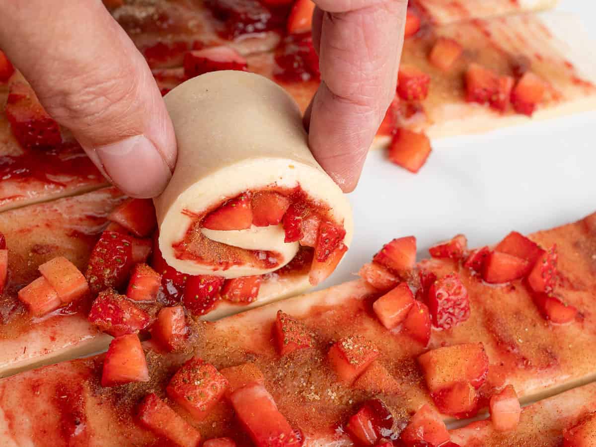 A hand rolls dough filled with chopped strawberries and cinnamon sugar, creating a spiral pastry reminiscent of homemade Strawberry Cinnamon Rolls. Other unrolled dough strips with fruit topping are visible on a white surface.