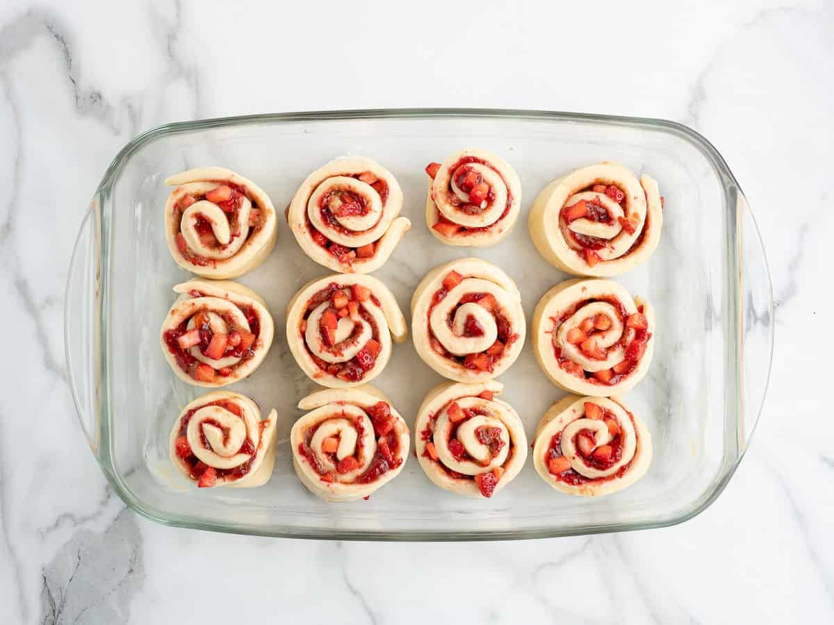 A glass baking dish with twelve unbaked Strawberry Cinnamon Rolls, showing spirals of dough and red fruit pieces, sits on a white marble surface.