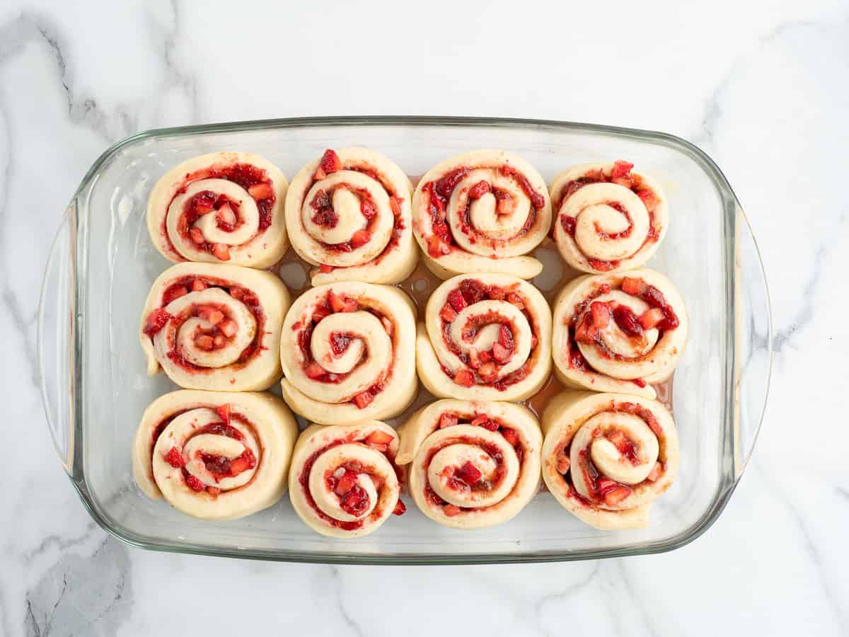 A glass baking dish with twelve unbaked Strawberry Cinnamon Rolls arranged in rows, sitting on a white marble surface.