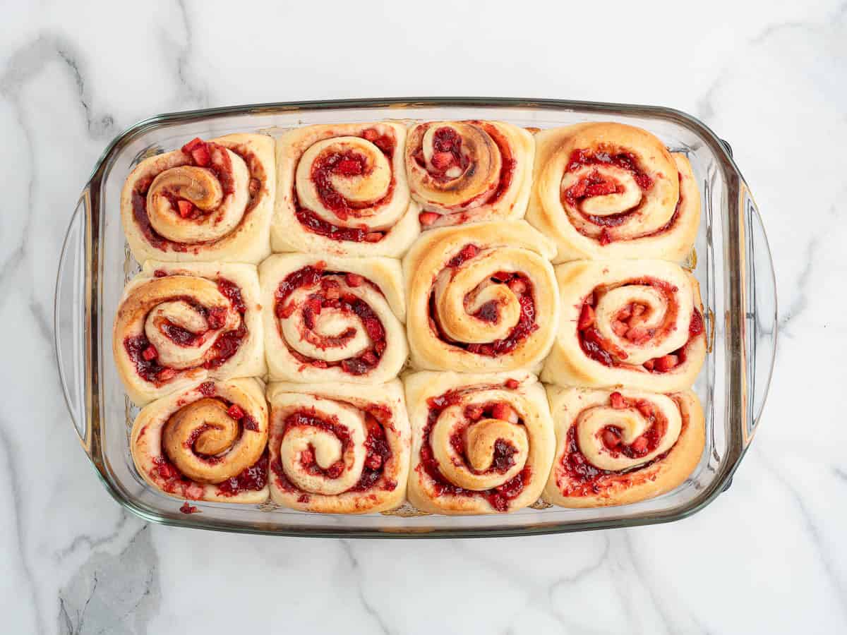 A glass baking dish filled with twelve golden, spiral-shaped Strawberry Cinnamon Rolls with a red fruit filling, arranged neatly on a white marble surface.