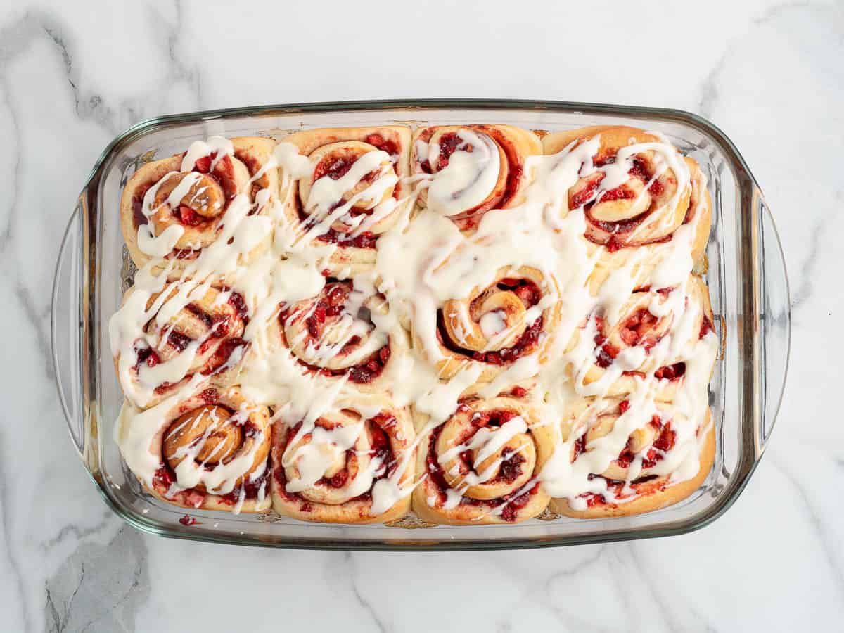 A glass baking dish filled with twelve spiral sweet rolls, reminiscent of Strawberry Cinnamon Rolls, topped with white icing, sits on a marble countertop.