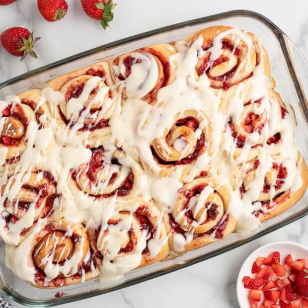A glass baking dish filled with Strawberry Cinnamon Rolls topped with white icing, surrounded by fresh strawberries and a bowl of diced strawberries on a marble surface.
