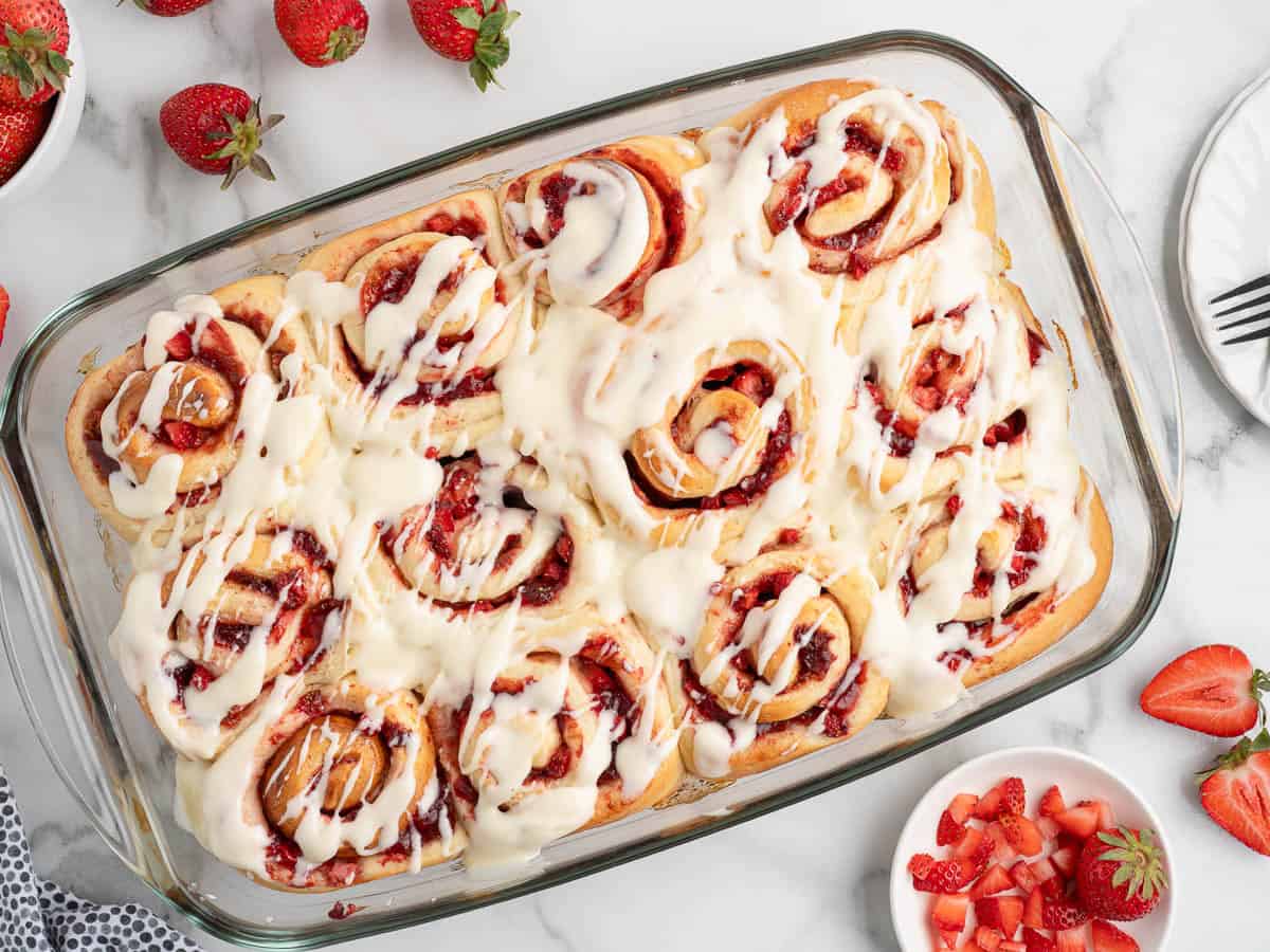A glass baking dish filled with Strawberry Cinnamon Rolls topped with white icing, surrounded by fresh strawberries and a bowl of diced strawberries on a marble surface.
