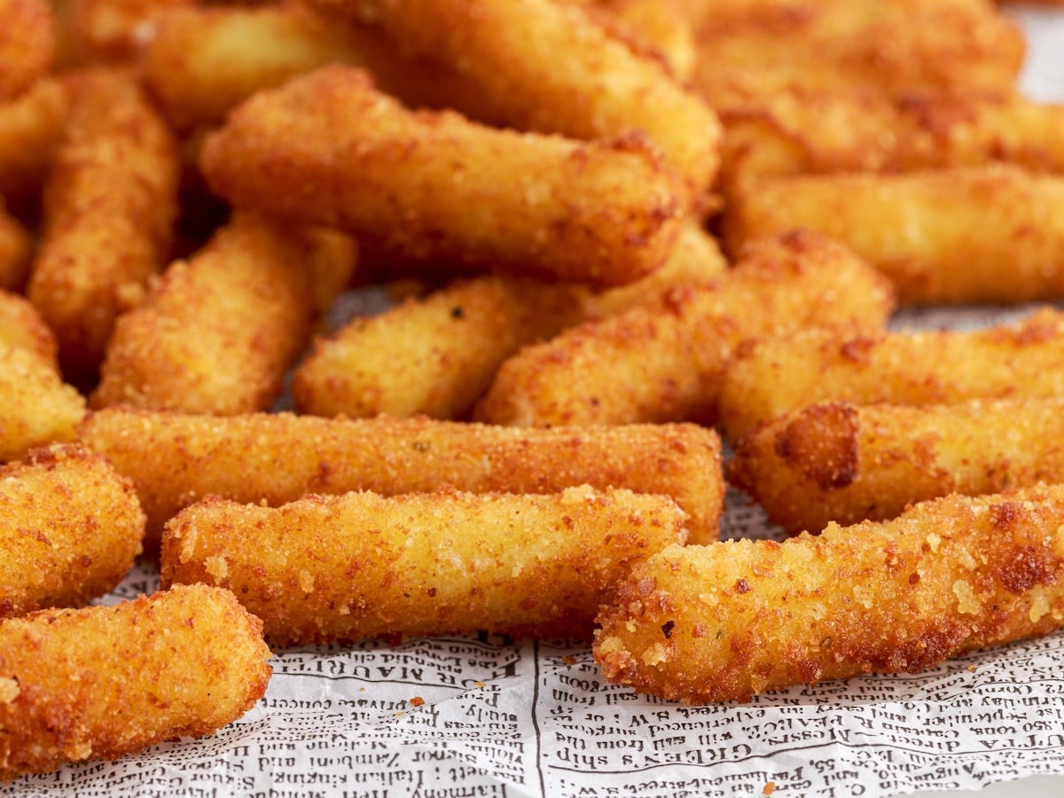 A close-up of crispy, golden-brown Stuffed Ziti Fritta sticks arranged on newspaper-style paper. The sticks appear freshly fried with a crunchy texture.