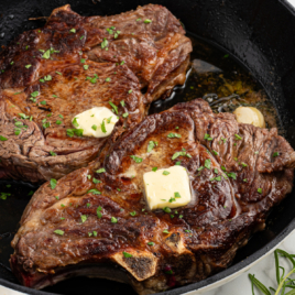 Two ribeye steaks are cooked in a skillet with butter melting on top of them.