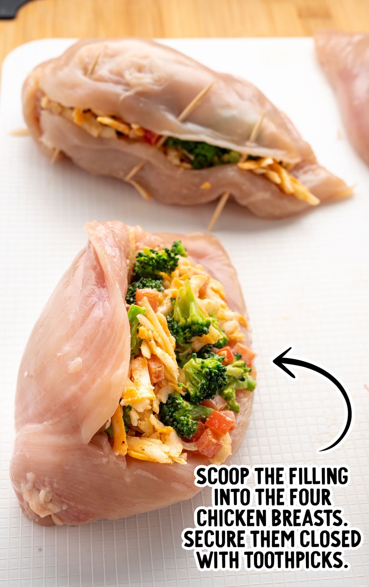 Close-up of raw chicken breasts stuffed with a broccoli and cheese mixture, secured with toothpicks, with text overlay instructing to scoop filling and secure with toothpicks.