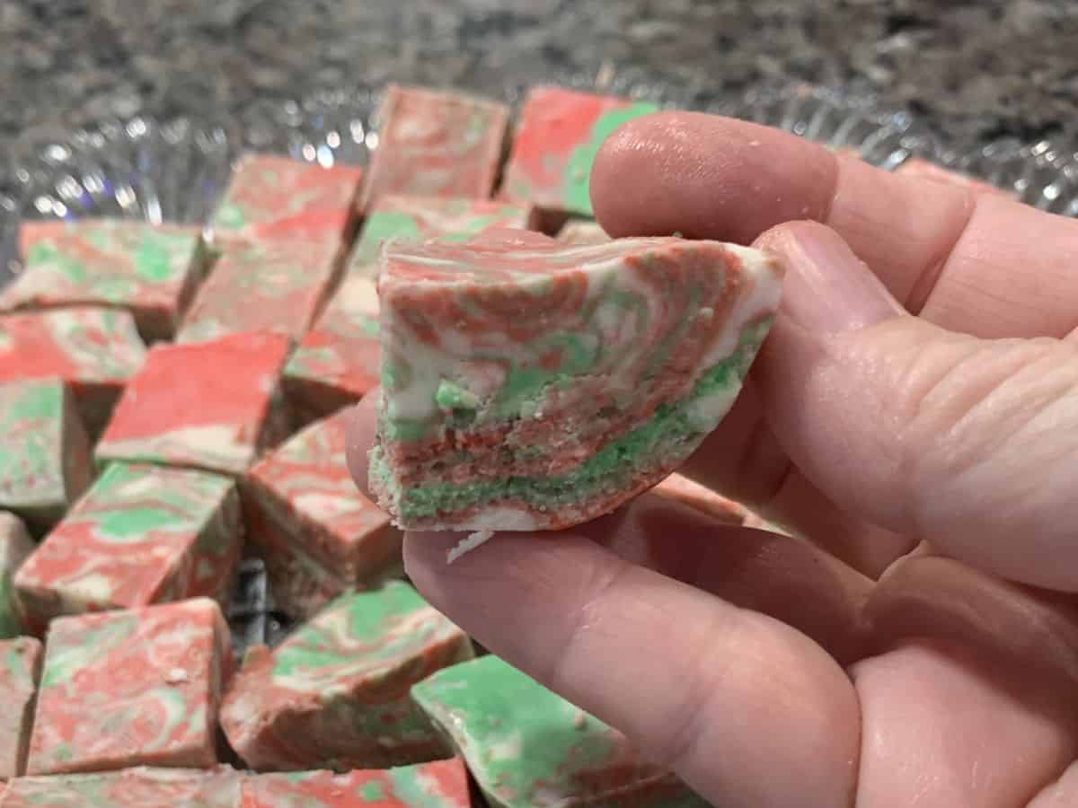 A hand holding a piece of red, green and white fudge made with frosting over a plate of more pieces.